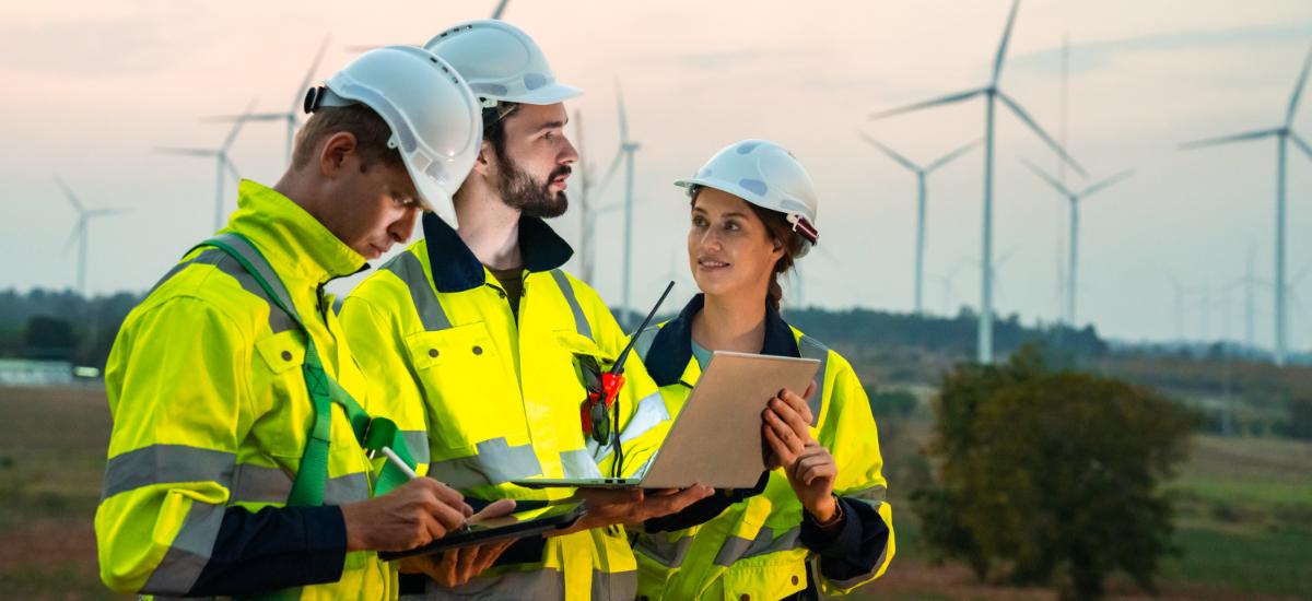 Three engineers in safety gear communicating via radio while reviewing laptop at wind farm.
